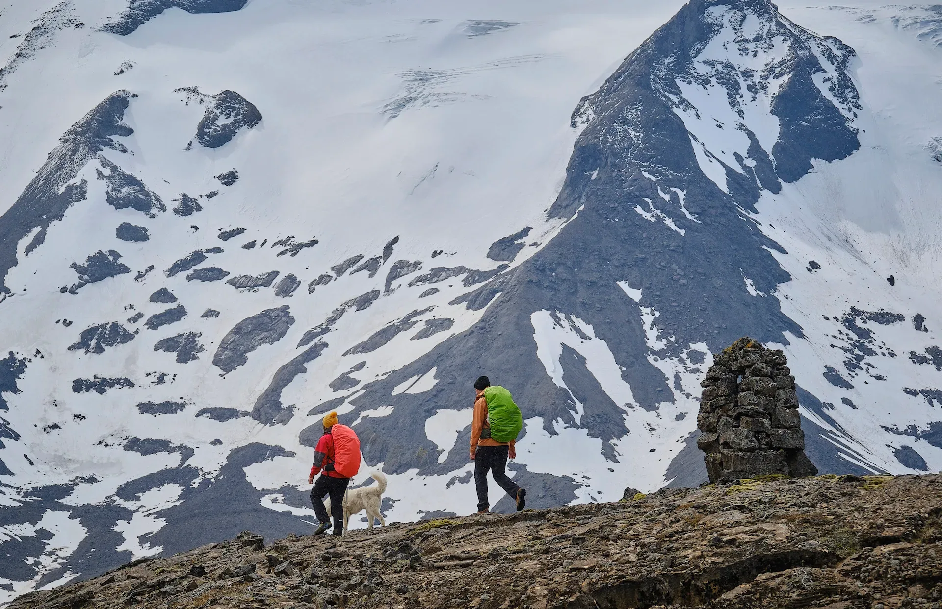Two men and a white siberian husky dog (Fenris the White Wolf) hiking past an ancient cairn, built by Vikings a thousand years ago. White mountain and glacier in the background.